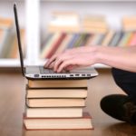 person typing on laptop using a pile of books as a table